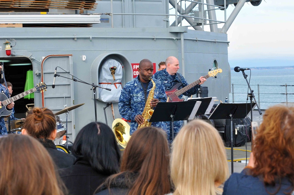 A military band is performing a live concert on the deck of a naval vessel, with musicians dressed in U.S. Navy blue digital camouflage uniforms. The performance includes at least a saxophonist, bassist, guitarist, and drummer playing for a crowd of civilian and military spectators. The setting appears to be a homecoming or public outreach event aboard a warship, with open water visible in the background.