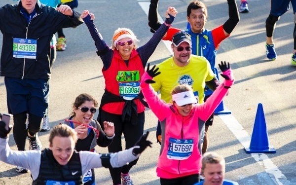 A group of runners participating in what appears to be the New York City Marathon, celebrating with raised arms and smiles. Multiple runners are visible wearing numbered race bibs, running gear, and cold-weather clothing. The scene captures a joyful, high-energy moment during the race on a paved road course.