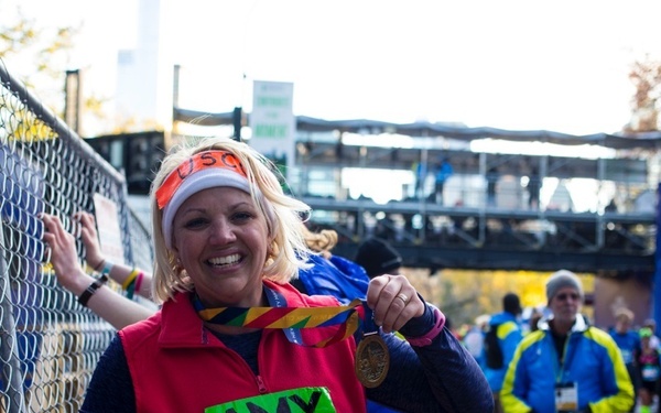 A woman named Amy, wearing bib number 69884, celebrates completing what appears to be the New York City Marathon. She is holding up a finisher's medal and smiling broadly while leaning against a chain-link fence. Other runners and spectators are visible in the background, and autumn foliage suggests a fall race date.