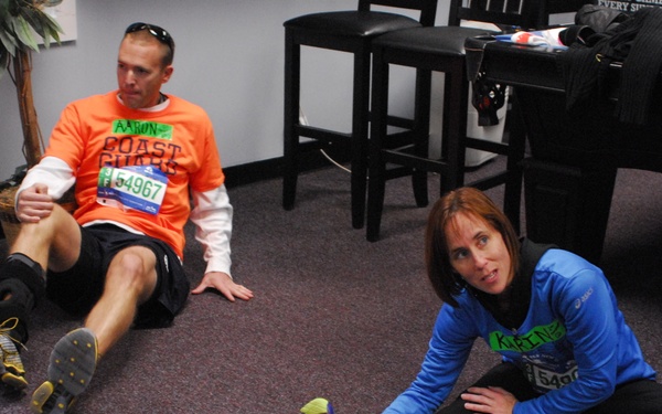 Two runners are sitting on a carpeted floor, likely resting or stretching after a race. The man in an orange 'Coast Guard' shirt wears bib number 54967, and the woman in a blue ASICS shirt wears bib number 5490-something. Both are wearing running shoes and appear to be in a recovery or warm-down phase indoors.