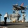 A U.S. Navy F-35C Lightning II fighter jet is captured in the process of landing or hovering above the flight deck of an aircraft carrier. Two personnel wearing white vests labeled 'Lightning Paddles' are observing the aircraft from the deck. The carrier's island superstructure is visible in the background with the number '68' displayed on it.