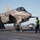 An F-35C Lightning II stealth fighter jet, numbered '75' with tail code 'VX-23', is positioned on the flight deck of a US Navy aircraft carrier. Ground crew members in color-coded jerseys are performing pre-flight or post-flight checks around the aircraft. The image was taken in clear daylight conditions with the ocean visible in the background.