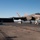 An F-35C Lightning II carrier-based stealth fighter jet is captured mid-launch or landing on what appears to be a US Navy aircraft carrier flight deck. The aircraft is in motion, evidenced by motion blur, with deck crew personnel visible in the background near a jet blast deflector. The scene takes place under clear blue skies over open ocean.