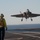 A fifth-generation stealth fighter jet, consistent with an F-35 variant, is captured in the final approach phase of a carrier landing on what appears to be an aircraft carrier flight deck. A deck crew member wearing a yellow jersey and helmet stands on the deck observing the aircraft. The ocean horizon is visible in the background under clear sky conditions.