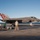 An F-35C Lightning II stealth fighter jet is captured during a carrier landing or takeoff on what appears to be a U.S. Navy aircraft carrier flight deck. The aircraft bears the number '75' and 'STRIKE TEST' markings, indicating it is a test aircraft. Several flight deck personnel in various colored jerseys are visible around the aircraft, with the ocean horizon visible in the background.