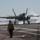 A fifth-generation stealth fighter jet, consistent with an F-35C, is landing on an aircraft carrier flight deck with arresting wire engagement visible, producing steam or smoke upon touchdown. A flight deck crew member wearing a yellow jersey marked 'FLT DK 302' observes the landing from a safe distance. The image captures a carrier arrested landing operation in what appears to be daylight conditions over open water.