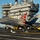 An F-35C Lightning II carrier variant jet aircraft is shown landing or taxiing on the flight deck of a U.S. Navy aircraft carrier. The aircraft bears the number '75' and markings reading 'STRIKE TEST' and 'NAVY', with a distinctive red and white checkered tail design. The carrier's island superstructure is prominently visible in the background under clear blue skies.