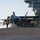 An F-35C Lightning II stealth fighter jet is positioned on the flight deck of a US Navy aircraft carrier, appearing to be in pre-launch or post-landing configuration with steam/exhaust visible near the landing gear. Multiple flight deck crew members in color-coded jerseys and helmets are visible surrounding the aircraft. The carrier's island superstructure is visible in the upper right background.