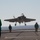 An F-35C Lightning II carrier variant fighter jet is captured moments before landing on an aircraft carrier flight deck, with its landing gear fully deployed. Several flight deck crew members in color-coded jerseys are positioned on the deck observing the approach. The ocean horizon is visible in the background, confirming the naval carrier operations setting.