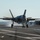An F-35C Lightning II fighter jet is captured during an arrested landing on what appears to be a naval aircraft carrier flight deck. The aircraft's tailhook has engaged the arresting wire, visible stretching across the deck, with steam or smoke rising from the friction. Several deck crew personnel are visible in the background observing the landing operation.