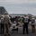 A group of naval personnel and observers stand on an aircraft carrier flight deck watching a jet aircraft launch at high speed, captured with significant motion blur. Personnel are wearing various colored vests and helmets indicating different roles including Safety Officer, PAO (Public Affairs Officer), FOC, and CVNP. The overcast sky and open ocean horizon confirm an at-sea carrier operations setting.