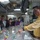 A man in a yellow sweater is conducting a briefing or demonstration at what appears to be an aircraft carrier's flight deck control station, using small colored aircraft models arranged on a tabletop display board. A group of approximately eight observers, appearing to be of East Asian and Western backgrounds, watches attentively. The setting is an interior shipboard compartment with monitors, control panels, and overhead equipment visible.