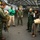 A U.S. Navy pilot in full flight gear appears to be conducting a briefing or demonstration for a group of visitors inside an aircraft carrier hangar bay. The group includes what appear to be Japanese military personnel and civilians, along with other U.S. Navy crew members. Military aircraft, including one marked '300', are visible in the background of the large hangar deck.