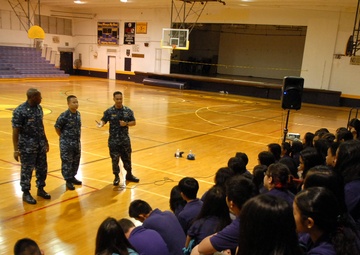 Four U.S. Navy personnel in blue digital camouflage uniforms are addressing a large group of students in a school gymnasium. The students, wearing purple uniforms, are seated in bleachers listening to what appears to be a military outreach or recruitment presentation. One sailor appears to be speaking to the audience while the others stand in formation beside him.