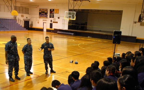 Four U.S. Navy personnel in blue digital camouflage uniforms are addressing a large group of students in a school gymnasium. The students, wearing purple uniforms, are seated in bleachers listening to what appears to be a military outreach or recruitment presentation. One sailor appears to be speaking to the audience while the others stand in formation beside him.