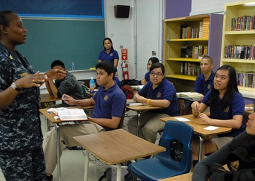 A U.S. Navy service member in digital camouflage uniform is addressing a group of students in a school classroom. The students, wearing matching blue polo uniforms, are seated at individual desks and appear attentive. A teacher or school staff member stands in the background near a chalkboard.