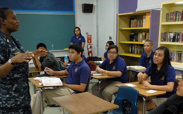 A U.S. Navy service member in digital camouflage uniform is addressing a group of students in a school classroom. The students, wearing matching blue polo uniforms, are seated at individual desks and appear attentive. A teacher or school staff member stands in the background near a chalkboard.