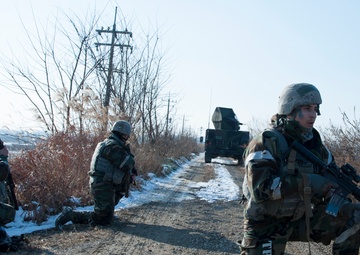 The image shows military personnel conducting what appears to be a training exercise along a rural dirt road in winter conditions with light snow on the ground. Three soldiers are visible in the foreground taking tactical positions, while a military vehicle is visible in the background on the road. The text 'TRNG ONLY' visible on one soldier's uniform confirms this is a training exercise rather than a combat operation.