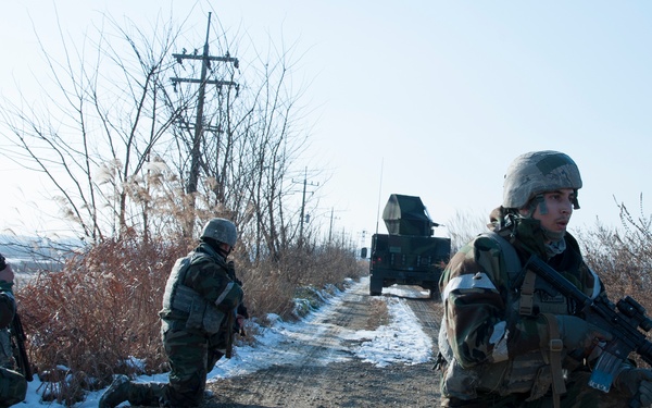 The image shows military personnel conducting what appears to be a training exercise along a rural dirt road in winter conditions with light snow on the ground. Three soldiers are visible in the foreground taking tactical positions, while a military vehicle is visible in the background on the road. The text 'TRNG ONLY' visible on one soldier's uniform confirms this is a training exercise rather than a combat operation.