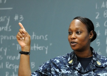 A U.S. Navy service member named Winfrey is standing in front of a chalkboard, appearing to teach or instruct a class. She is wearing Navy Working Uniform (NWU) digital camouflage and has one finger raised, suggesting she is making a point or explaining something. The chalkboard behind her contains vocabulary lesson content including word lists and a short story reference to 'The Lottery.'