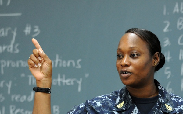A U.S. Navy service member named Winfrey is standing in front of a chalkboard, appearing to teach or instruct a class. She is wearing Navy Working Uniform (NWU) digital camouflage and has one finger raised, suggesting she is making a point or explaining something. The chalkboard behind her contains vocabulary lesson content including word lists and a short story reference to 'The Lottery.'