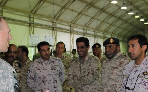 A US Army soldier in ACU (Army Combat Uniform) digital camouflage is briefing or speaking to a group of military personnel wearing what appear to be Gulf state armed forces uniforms in desert camouflage patterns. The meeting is taking place inside a large military tent or hangar facility. The group appears to be engaged in a multinational military exchange or coordination meeting.