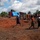The image shows a group of people gathered on a cleared, red-soil open area in what appears to be a rural African setting. A blue tarpaulin structure and a black water tank are visible in the background, suggesting a temporary or emergency setup. The scene depicts community activity with multiple individuals of varying ages moving across the open ground.