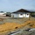 The image shows an active construction or installation site, likely a temporary military or humanitarian camp. Several structures including white modular buildings, military-style tents, and a wooden-framed covered shelter with a metal roof are visible. The site appears to be under active development, with excavated trenches, lumber, and metal conduit pipes in the foreground.