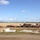 The image shows an open, flat construction or staging area with large quantities of lumber/timber stacked in multiple piles. Several workers are visible near the timber stacks, with at least one wearing a high-visibility orange vest. The setting appears to be a wide, cleared land area — possibly an airfield or large development site — under a partly cloudy blue sky.