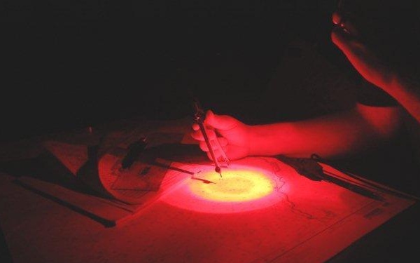 A person is working on a nautical or navigational chart in near-total darkness, illuminated by a red light source. They are using a drawing compass (dividers) to measure or plot a position on the chart. The red lighting is consistent with night navigation practices used aboard ships or in military/aviation settings to preserve night vision.