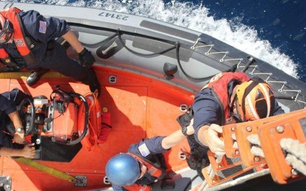 An aerial/overhead view of what appears to be a US Coast Guard rigid inflatable boat (RIB) with personnel conducting operations alongside a larger vessel. At least three crew members are visible, with one climbing or managing a rope ladder connecting the small boat to the larger vessel. The boat hull bears the number '23147' and personnel are wearing standard Coast Guard gear including helmets and life vests.