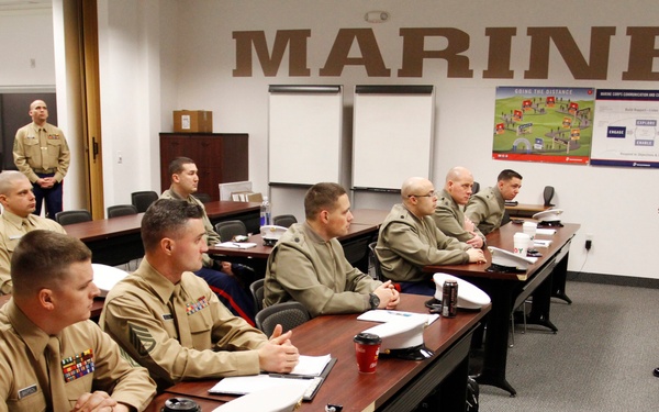 A group of United States Marines in uniform are attending a classroom briefing or training session. One Marine in dress blues with red stripe trousers is standing at the front, apparently presenting or instructing the seated attendees. The room is clearly identified as a Marine Corps facility by the large 'MARINES' lettering on the wall.