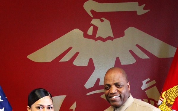 Two U.S. Marine Corps personnel in service uniforms are shaking hands in front of a red wall bearing the Marine Corps Eagle, Globe, and Anchor emblem. An American flag and a Marine Corps flag are visible in the background. The occasion appears to be a formal recognition or promotion ceremony.
