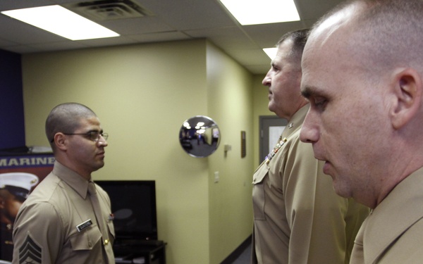 A formal military ceremony is taking place in what appears to be a Marine Corps recruiting office or administrative space. A senior Marine officer is reading from an official certificate or commendation document held in a red presentation folder, while a lower-ranked Marine (Sergeant) stands at attention facing him. Two officers are present on the right side, with one prominently displaying service ribbons on his uniform.