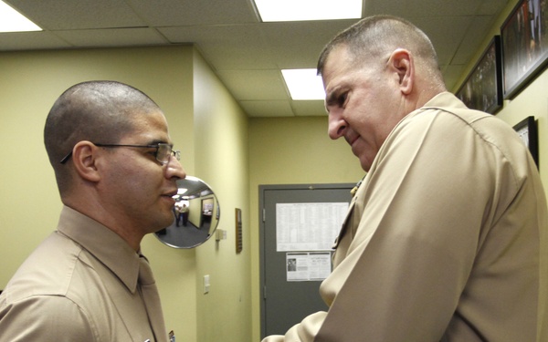 Two U.S. Marine Corps personnel are engaged in what appears to be a formal recognition or pinning ceremony inside an office or administrative building. A senior officer is presenting or pinning something onto the chest of a Sergeant, who is wearing glasses and displaying chevron rank insignia on his sleeve. The setting appears to be a recruiting or administrative office environment.