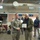 A military ceremony taking place in what appears to be a transit or rest stop facility with a Starbucks coffee shop visible in the background. A soldier in Army Combat Uniform is shaking hands with a civilian man in a green jacket while both hold a certificate or plaque, suggesting an award or recognition presentation. Several other soldiers and civilians are present as witnesses in the background.