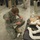 A U.S. Army soldier in ACU (Army Combat Uniform) is kneeling on a concrete floor, working with PVC pipe components and wiring. The setting appears to be an indoor facility, possibly a training or demonstration environment, with multiple other military and civilian personnel observing in the background. The soldier appears to be assembling or inspecting a device made from PVC pipes and electrical components.
