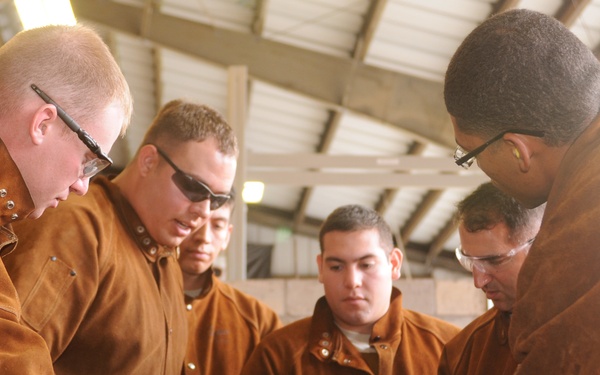 A group of five individuals wearing brown leather welding jackets and safety glasses are gathered around a workbench in an industrial or training facility. They appear to be reviewing documents or plans laid flat on the surface, suggesting a instructional or briefing session. The setting appears to be a workshop or training bay with a metal roof structure visible in the background.
