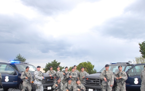 A group of approximately 16 military personnel in US Air Force Airman Battle Uniform (ABU) camouflage pattern pose for a group photo in a parking lot. They are armed with rifles and wearing black berets, positioned in front of two dark navy blue Security Forces patrol vehicles. The vehicles display blue emergency lights and Security Forces shield emblems on the doors.