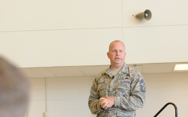 A senior U.S. Air Force enlisted member, identifiable by Chief Master Sergeant rank insignia, stands at the front of a room addressing a seated audience of uniformed military personnel. The speaker appears to be conducting a briefing or all-call meeting in what looks like a military facility. The audience members are seated with their backs to the camera, listening attentively.