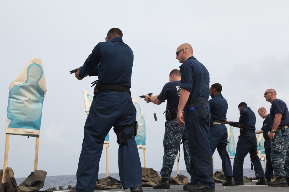 A group of military or law enforcement personnel in navy blue uniforms are conducting a firearms qualification or training exercise on what appears to be the deck of a naval vessel. Multiple individuals are aiming handguns at human-silhouette paper targets mounted on easel stands. The overcast sky and visible ocean horizon confirm an open-water shipboard setting.