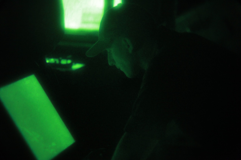 A person wearing a cap is visible in a very dark environment illuminated primarily by green-tinted light sources, likely screens or monitors. The scene appears to be a dimly lit operations room, cockpit, or command station with multiple glowing green displays. The monochromatic green lighting suggests possible use of night-vision photography or red-light-restricted environment with green instrument lighting.