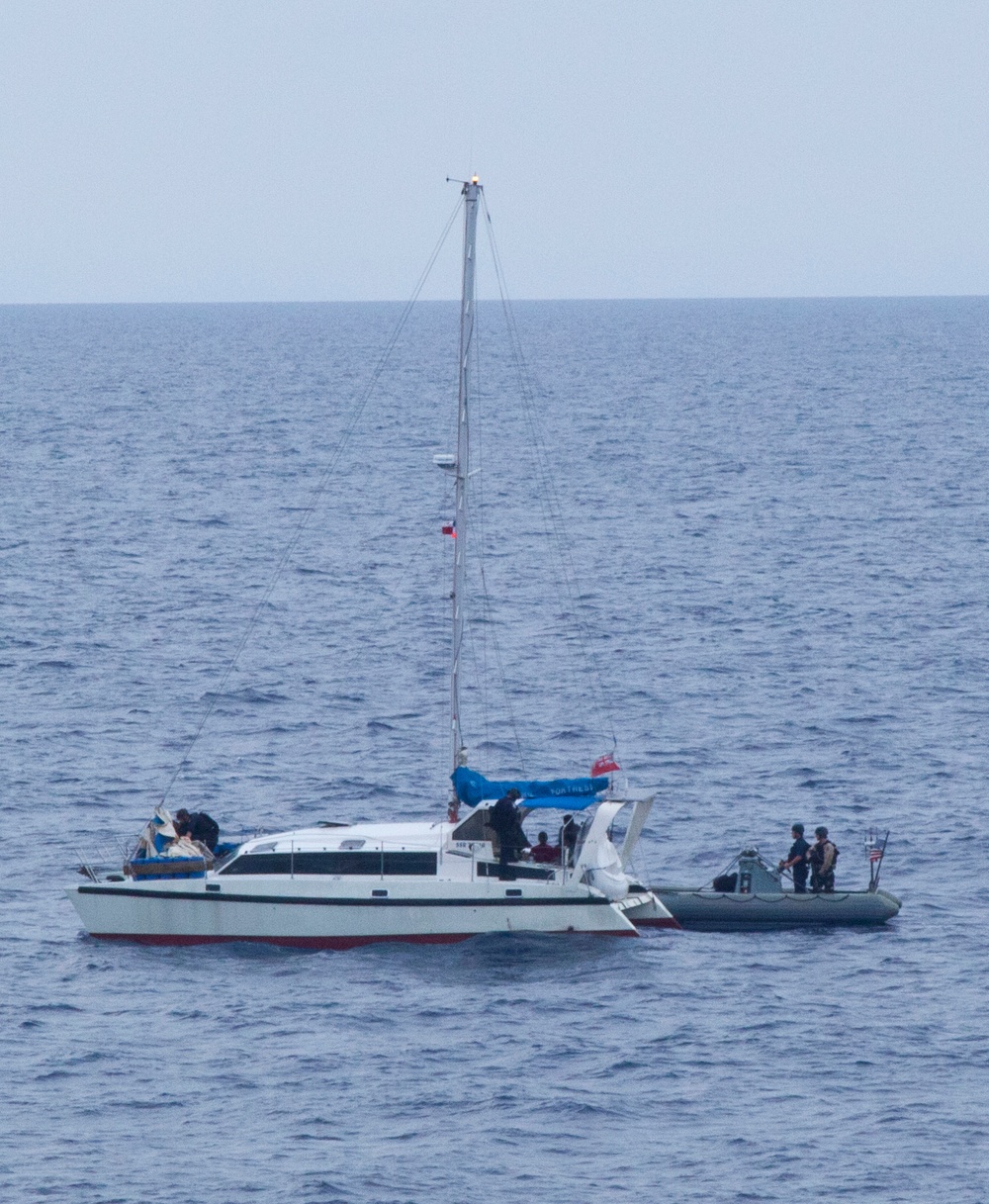 A white catamaran sailboat is stopped at sea with its sails furled and a blue tarp visible on deck. A gray rigid inflatable boat (RIB) with several personnel aboard is positioned alongside the starboard stern of the catamaran. The scene appears to depict a maritime law enforcement or coast guard boarding operation, with personnel visible on both vessels.