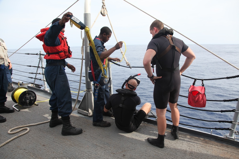 The image shows a group of personnel on the deck of a naval or coast guard vessel preparing for a diving operation. Two crew members in blue uniforms, one wearing an orange life vest, are handling rigging equipment including yellow straps and ropes attached to a mast. Two individuals in black wetsuits are also present, one seated on the deck and one standing, suggesting they are divers being prepared for water entry.