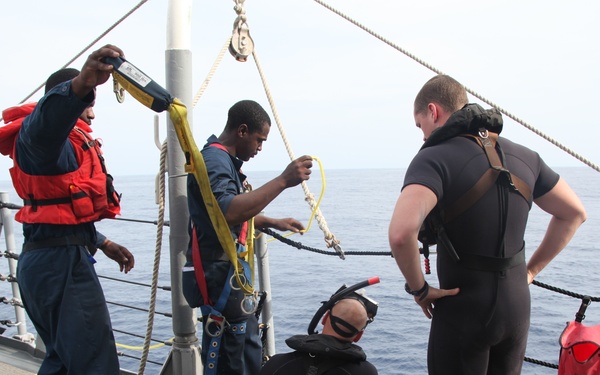The image shows a group of personnel on the deck of a naval or coast guard vessel preparing for a diving operation. Two crew members in blue uniforms, one wearing an orange life vest, are handling rigging equipment including yellow straps and ropes attached to a mast. Two individuals in black wetsuits are also present, one seated on the deck and one standing, suggesting they are divers being prepared for water entry.