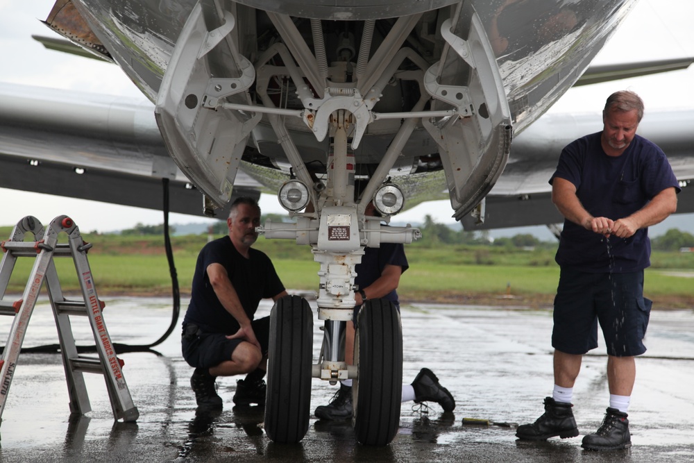 Three maintenance technicians are working on the nose landing gear of a large aircraft on a wet tarmac. The workers, dressed in dark navy uniforms, are inspecting and servicing the nose gear assembly, which includes dual wheels, struts, and landing lights. A folding ladder is visible to the left, and the surrounding environment suggests a tropical or humid airfield setting.