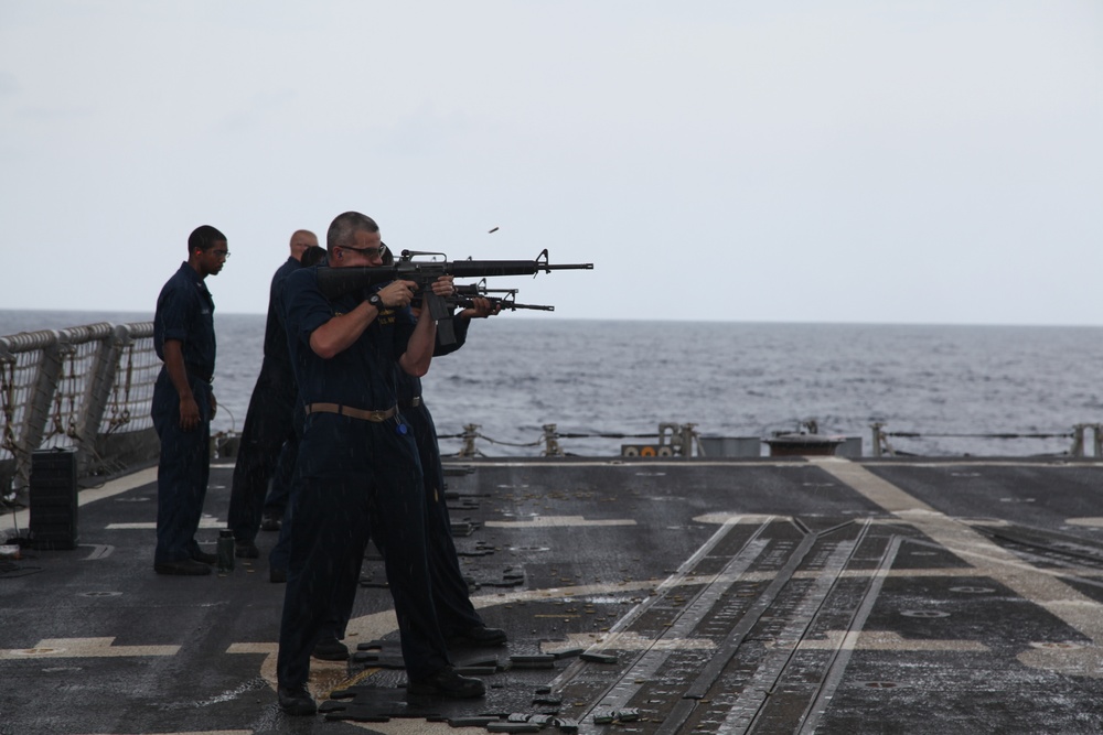 Military personnel are conducting a live-fire weapons training exercise on the deck of a naval vessel at sea. Two individuals in the foreground are actively firing M16-style rifles over the ship's railing toward the open ocean, while others stand nearby observing. A spent brass casing is visibly ejecting from one of the rifles.