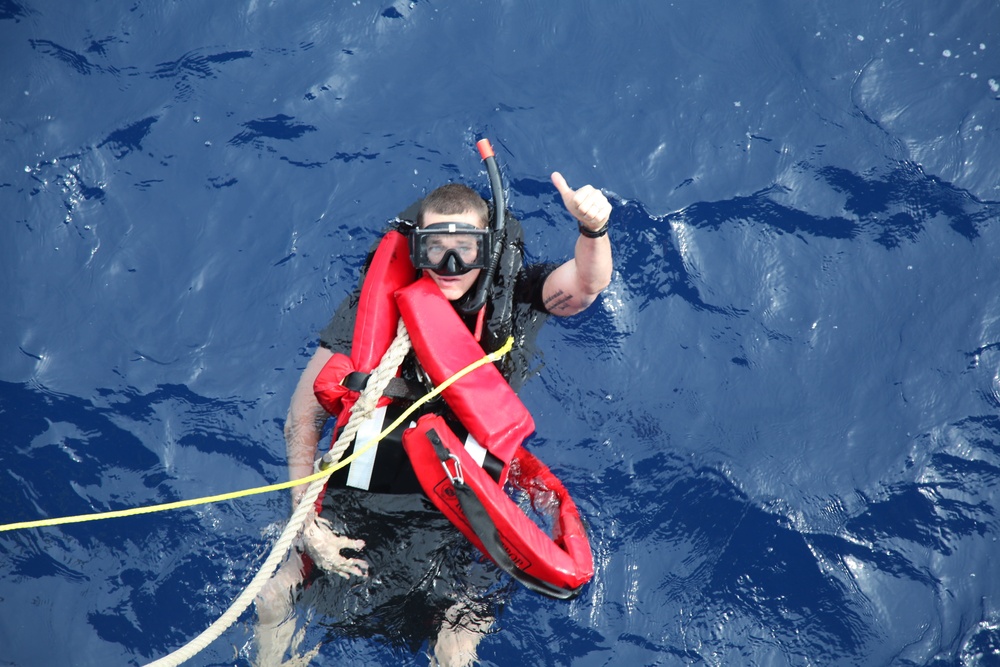 A person is floating in open blue ocean water wearing snorkeling gear and a red inflatable life preserver/rescue buoy. They are holding a white rope and a yellow line, and giving a thumbs-up gesture toward the camera. The image appears to depict a water rescue drill or safety training exercise.
