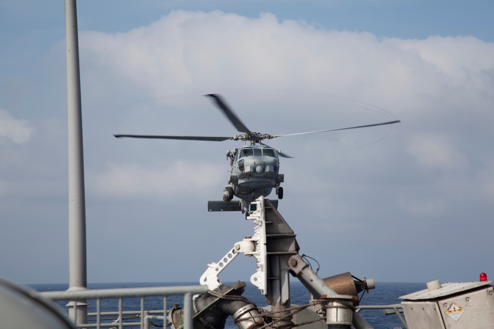 A military helicopter, consistent with an SH-60 Seahawk type, is hovering or approaching a naval vessel's deck area. The image is taken from the ship's deck, showing various shipboard equipment and structures in the foreground. The scene appears to depict a shipboard helicopter operation over open ocean.
