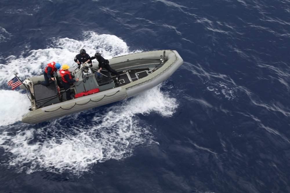An aerial view of a gray rigid inflatable boat (RIB) underway on open deep-blue ocean water. The vessel displays a small American flag at its stern and carries several personnel in various colored safety gear. The boat is moving at speed, generating a visible wake on both sides.